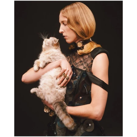 A woman in a dark patterned dress holding a light-colored cat against a black background, photographed by Aart Verrips.