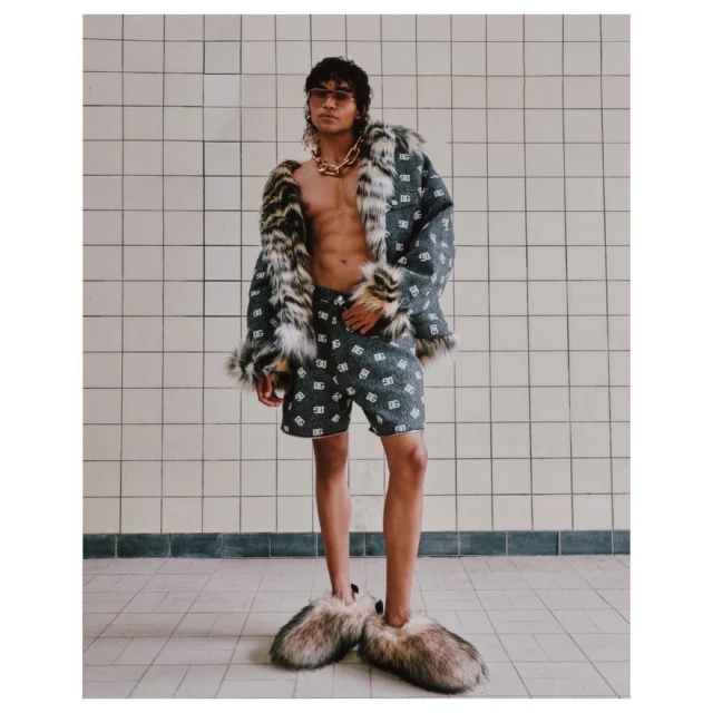 A man posing indoors wearing a patterned jacket with fur trim, matching shorts, sunglasses, and furry slippers, with a gold chain.