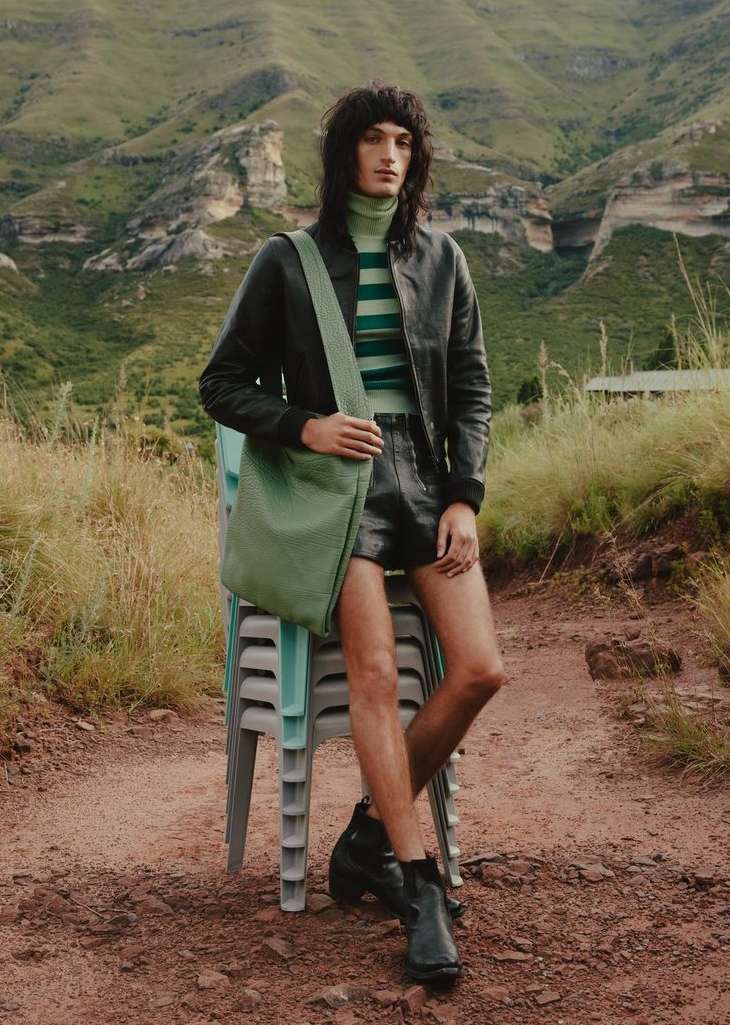 Man wearing leather jacket and shorts with messenger bag poses in a field with mountains. Fashion shot with natural light.