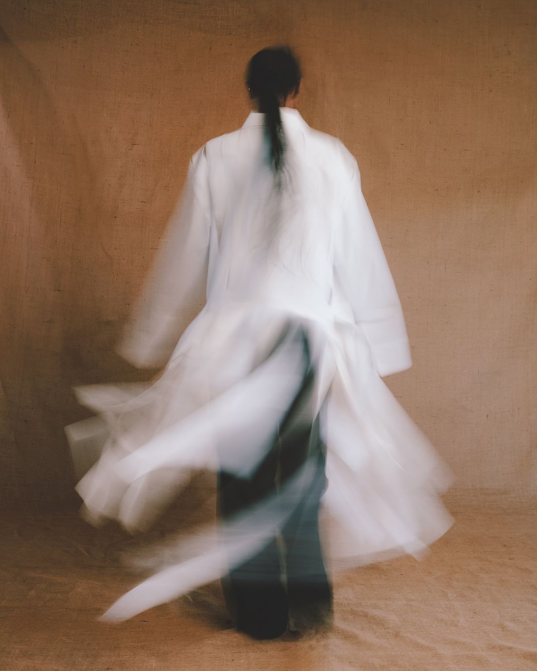 A person with dark hair seen from behind wearing a white coat and dark pants, captured with motion blur against a textured neutral background.