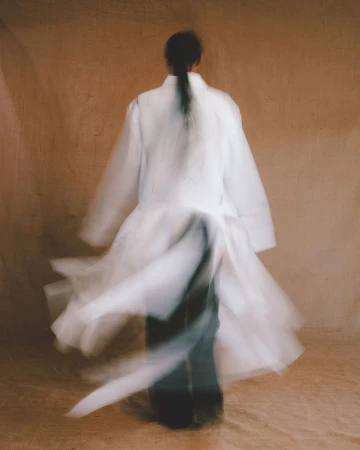 A person with dark hair seen from behind wearing a white coat and dark pants, captured with motion blur against a textured neutral background.