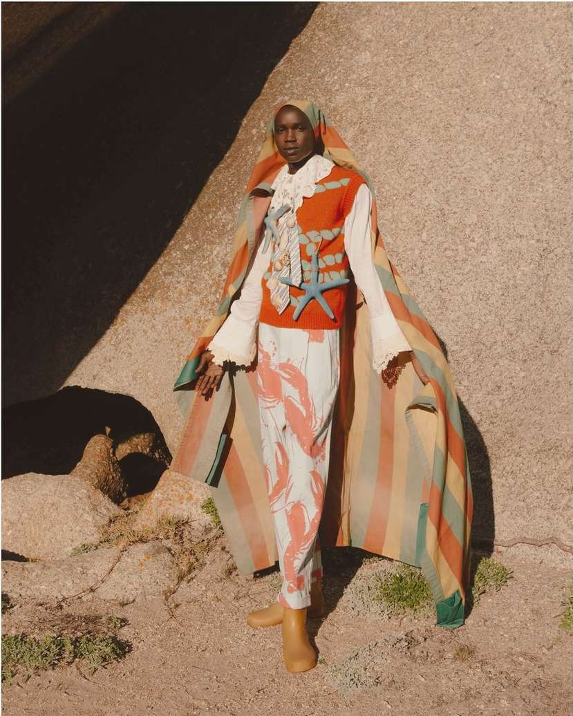 A fashion portrait features a Black model wearing a striking lobster-print outfit, set against a sun-drenched, rocky outdoor backdrop.