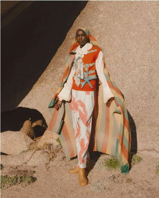A fashion portrait features a Black model wearing a striking lobster-print outfit, set against a sun-drenched, rocky outdoor backdrop.