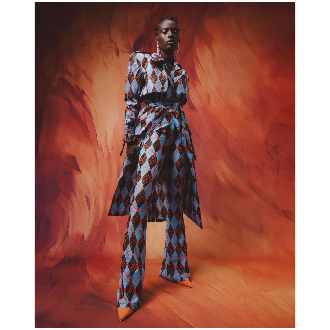 Full-length portrait of an African female model in a blue and brown diamond-patterned pantsuit, posing against an abstract orange background.