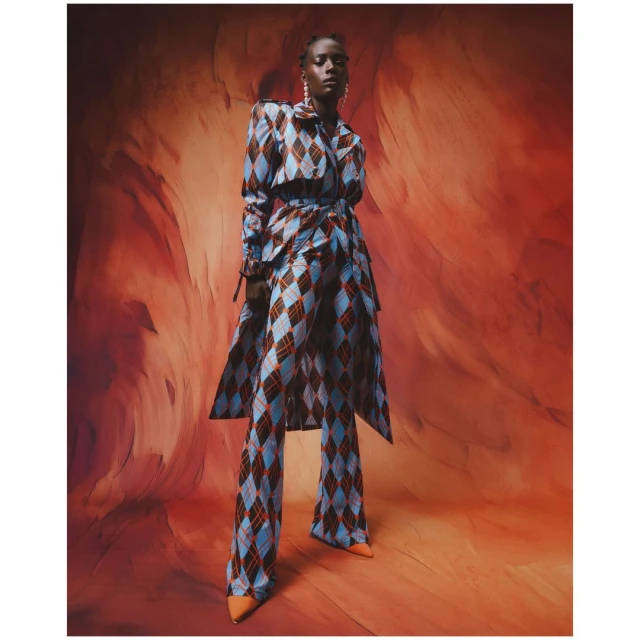 Full-length portrait of an African female model in a blue and brown diamond-patterned pantsuit, posing against an abstract orange background.