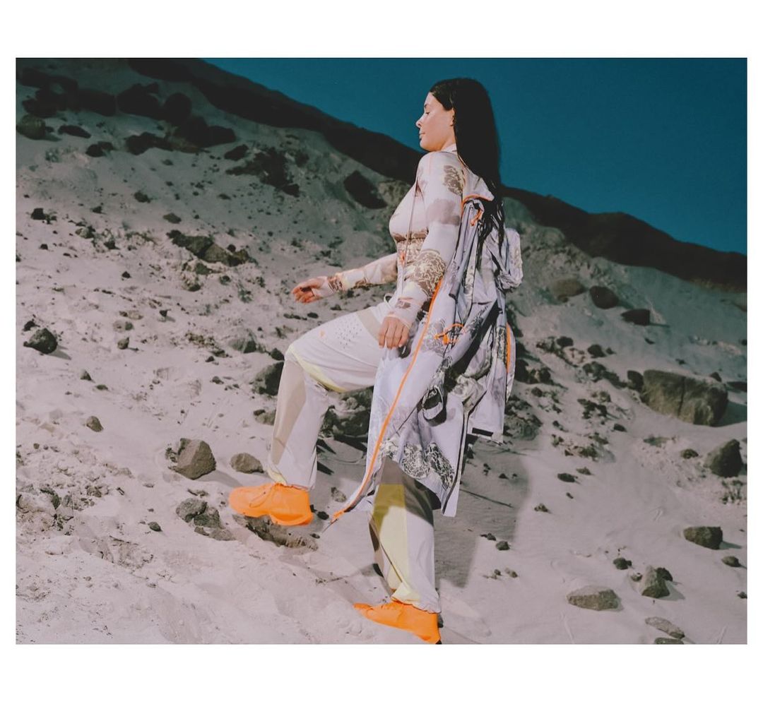 A woman in patterned clothing walks up a sandy hill wearing bright orange sneakers, with a printed jacket over her shoulder.