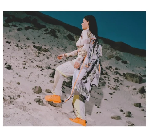 A woman in patterned clothing walks up a sandy hill wearing bright orange sneakers, with a printed jacket over her shoulder.