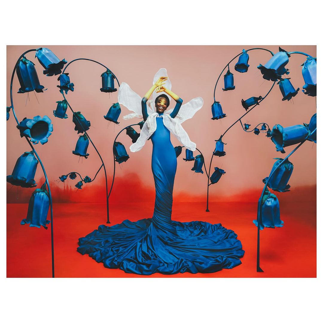 Elegant African-American woman posing in a blue dress with floral accents, set against a salmon and red backdrop, creating a dreamy fashion editorial.