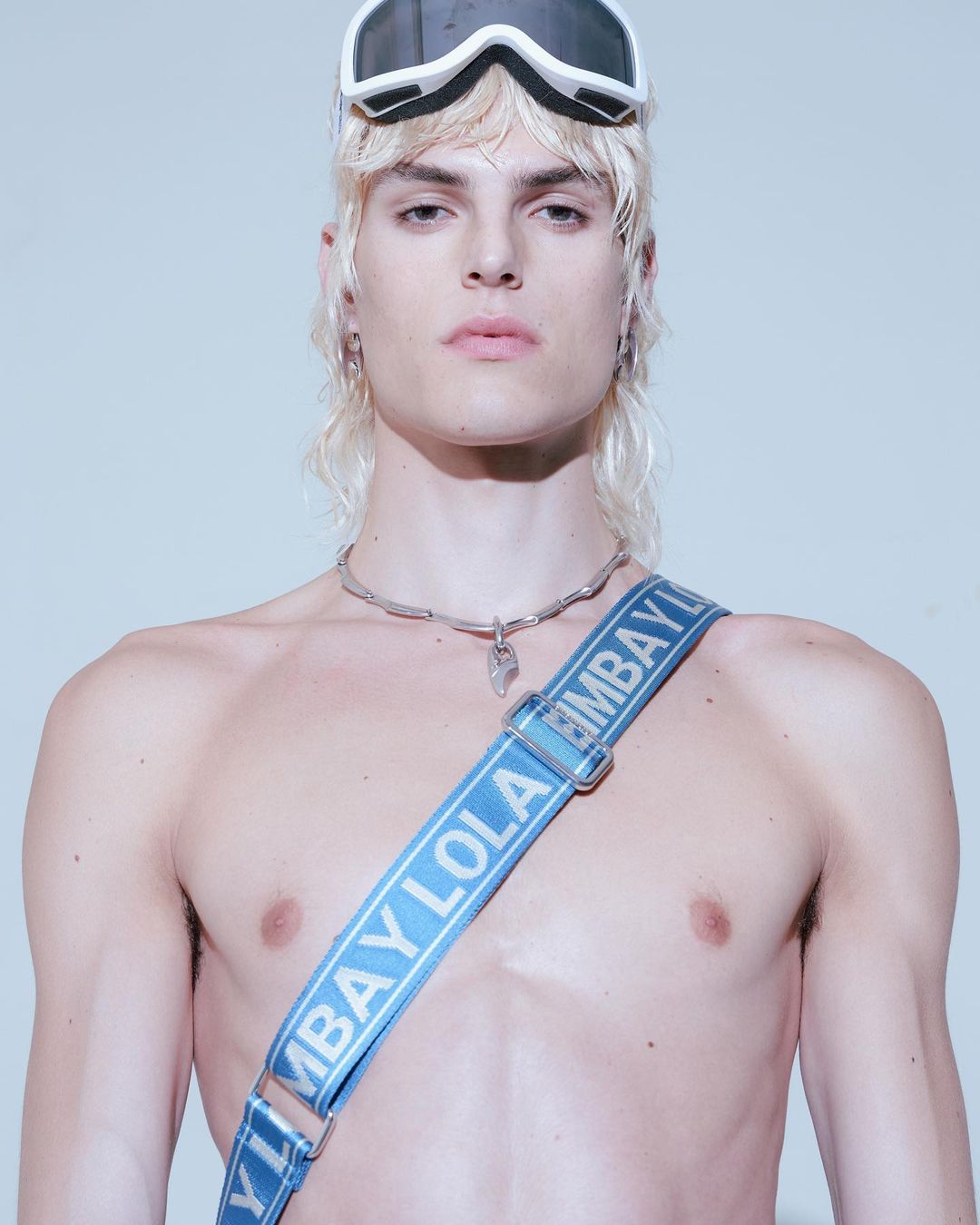 Young adult male model with platinum blonde mullet, sunglasses, and blue strap, posing indoors against a white background in a fashion shot.