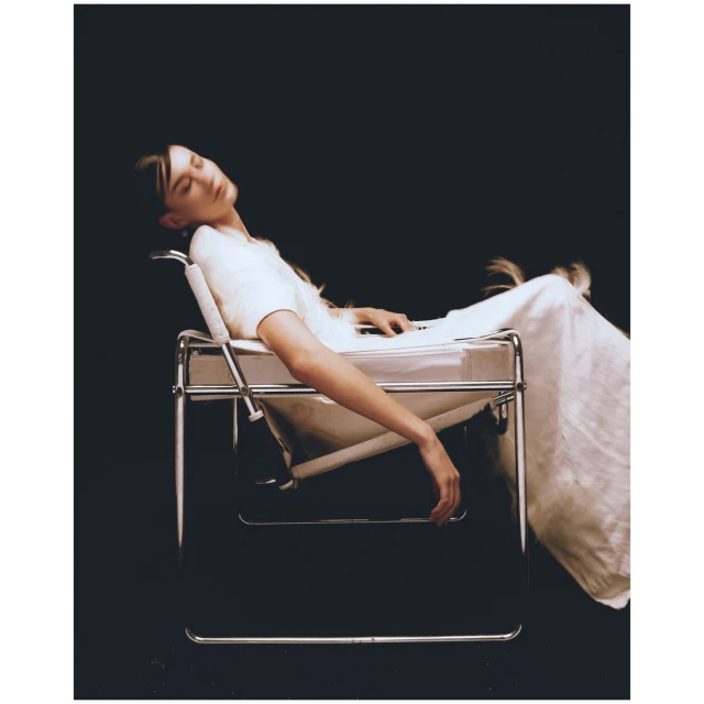 A serene image shows a young woman in a white dress reclining in a modernist chair against a dark backdrop in a studio setting.