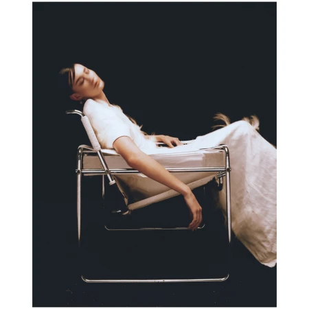 A serene image shows a young woman in a white dress reclining in a modernist chair against a dark backdrop in a studio setting.