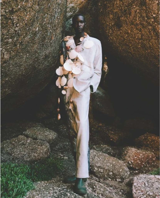 Fashionable dark-skinned male model posing in an off-white suit and shell adornments, standing among large stones and greenery outdoors.