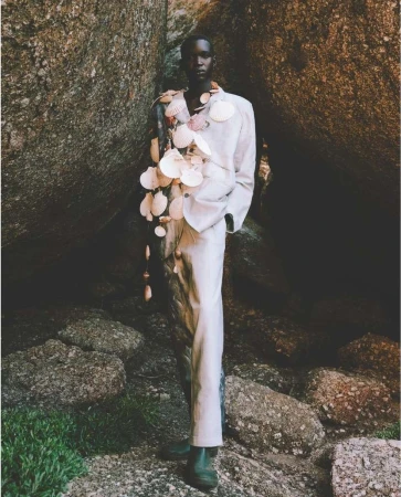 Fashionable dark-skinned male model posing in an off-white suit and shell adornments, standing among large stones and greenery outdoors.