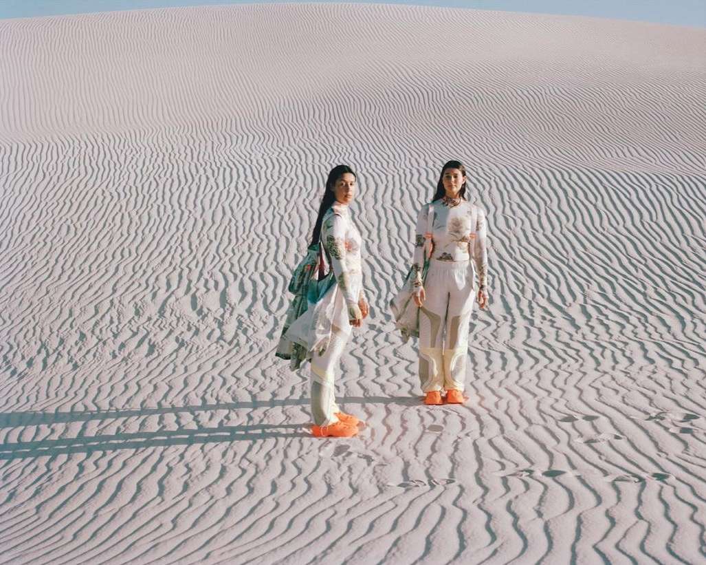 Two women stand on sand dunes in matching outfits with long sleeves and light colors.  The landscape shows the patterns of the rippled sand.