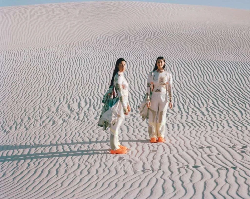 Two women stand on sand dunes in matching outfits with long sleeves and light colors.  The landscape shows the patterns of the rippled sand.