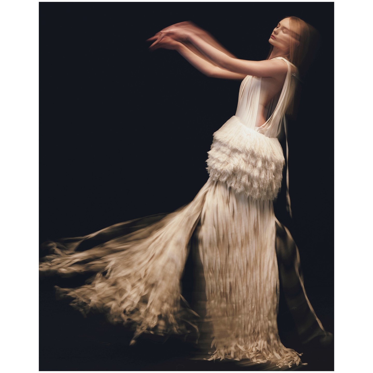 Woman in motion wearing a flowing, fringed white gown against a dark background, capturing the essence of movement and fashion.