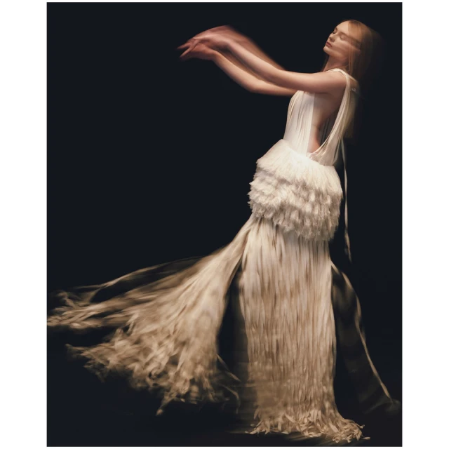 Woman in motion wearing a flowing, fringed white gown against a dark background, capturing the essence of movement and fashion.