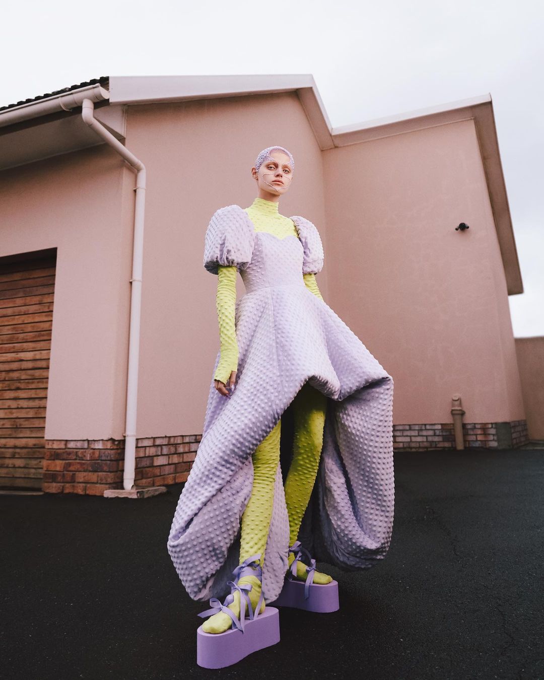 A model poses outdoors in a light purple dress with puffed sleeves, chartreuse top and leggings, and platform shoes in an editorial fashion shot.