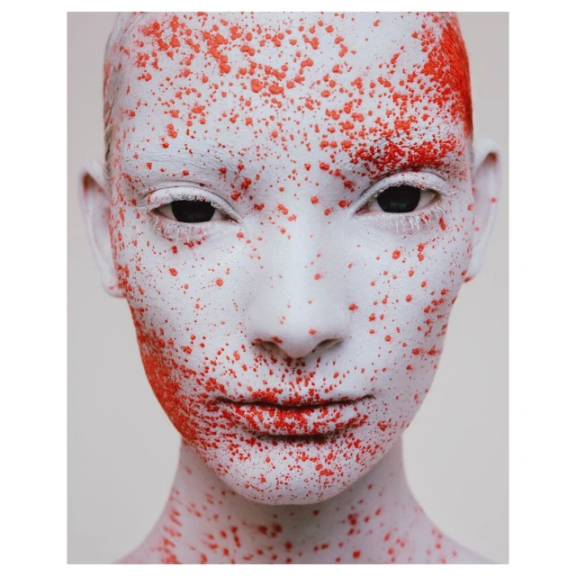 Artistic close-up portrait of a person with face and head covered in white makeup and dotted with bright orange specks, against a white backdrop.