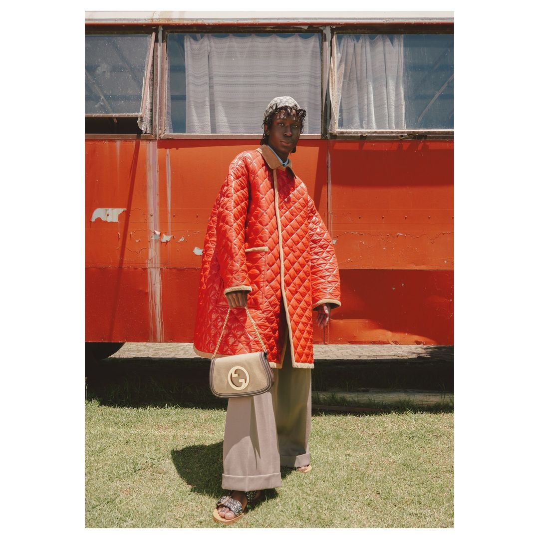 A Black model poses in front of a vibrant orange van, wearing a red quilted coat, tan wide-leg pants, and a GG logo crossbody bag.