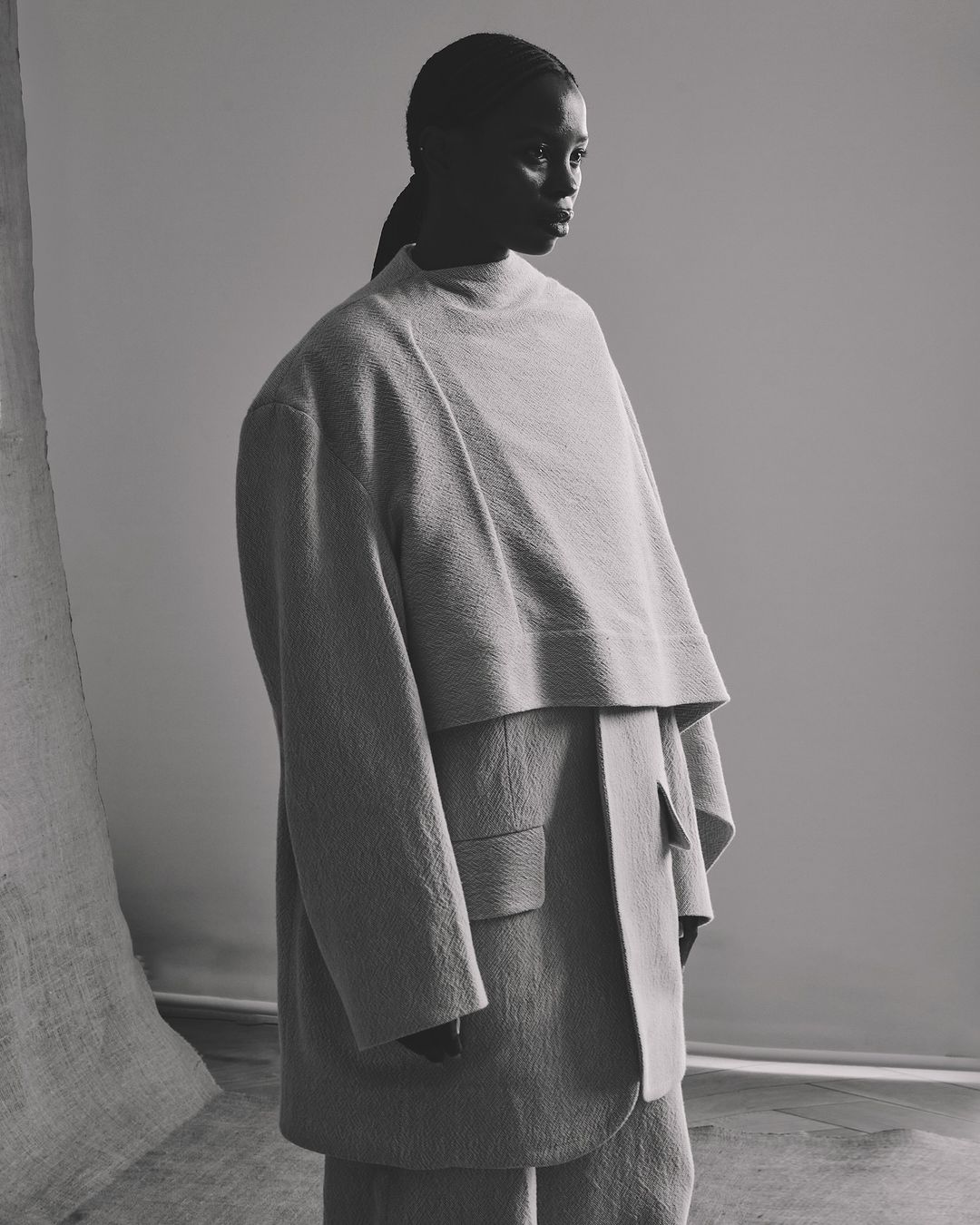 Monochrome portrait of a black woman in oversized beige suit, looking to the side in a studio setting with draped fabric.