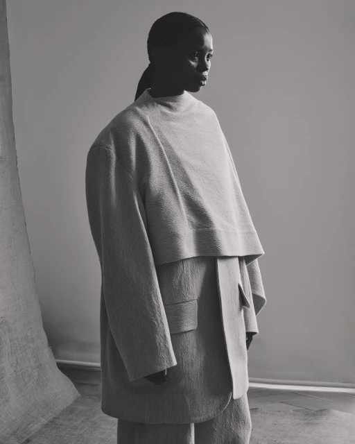 Monochrome portrait of a black woman in oversized beige suit, looking to the side in a studio setting with draped fabric.