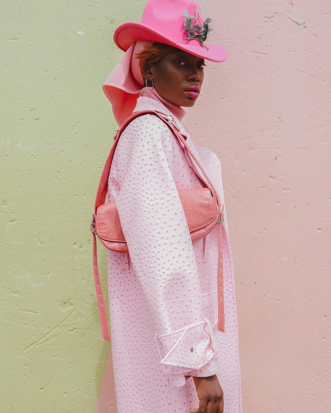 A woman poses in a pink hat, coat, and purse against a split pink-and-green wall, showcasing a coordinated fashion ensemble.