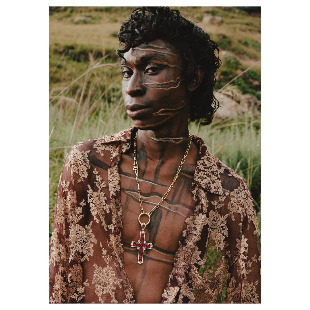 Close-up of a model with tribal face paint wearing a sheer floral shirt and gold cross necklace, posing outdoors with greenery.