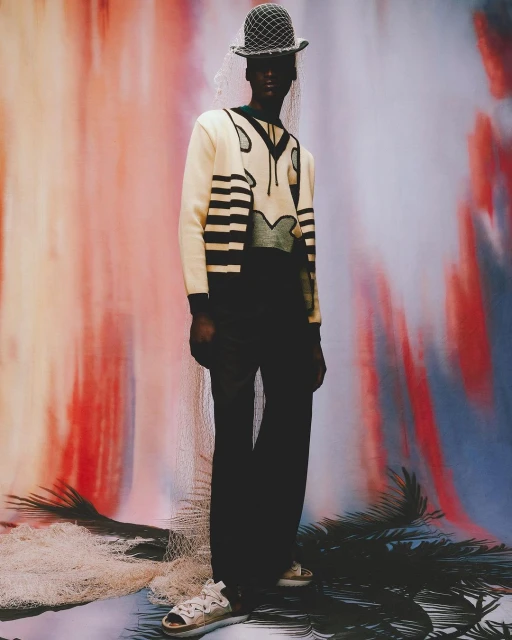 A dark-skinned model in V-neck sweater and wide-leg pants stands against an abstract painted backdrop wearing a net-veiled bucket hat.