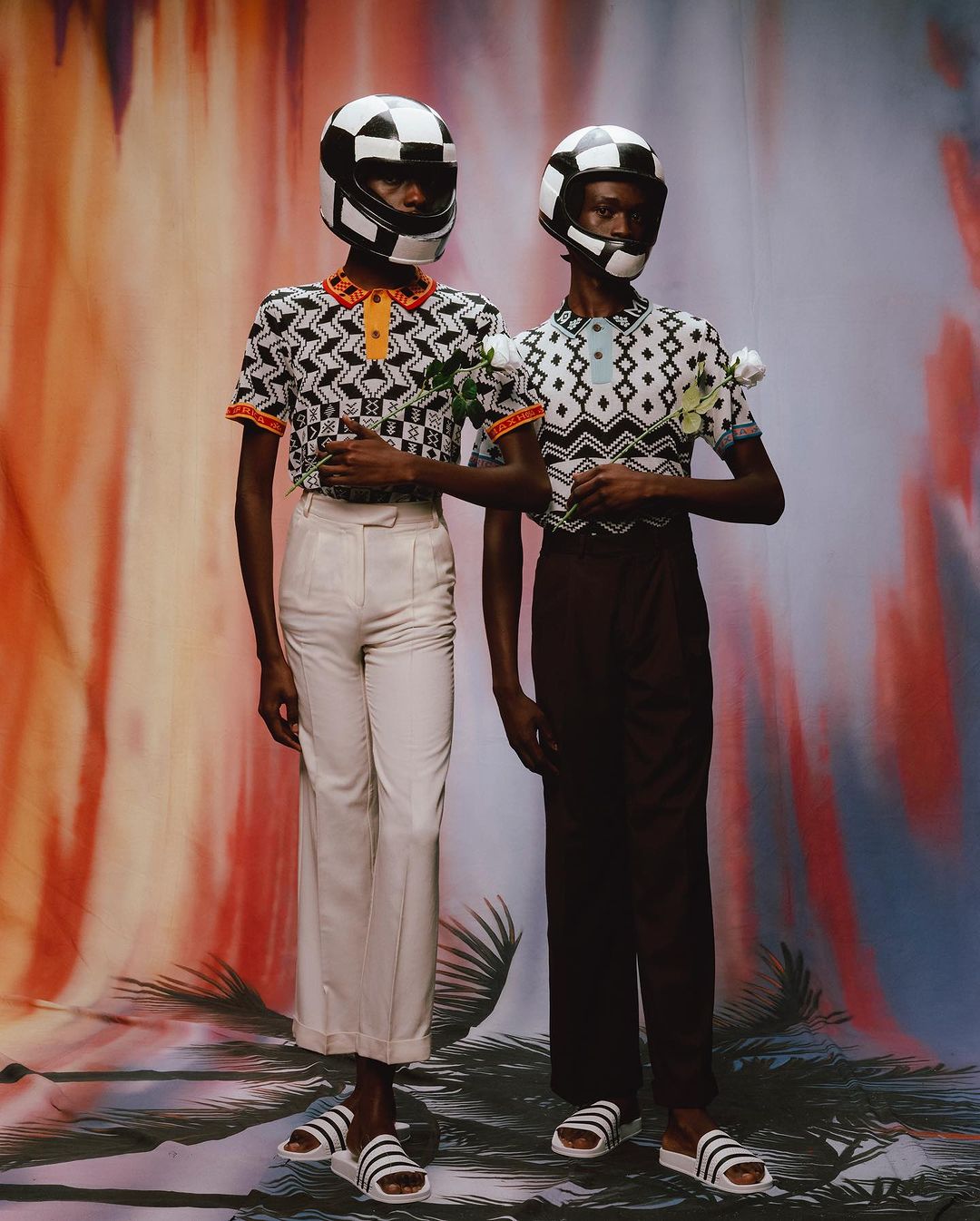 Two models wear geometric-print tops, cream and brown trousers, and checkered motorcycle helmets against a vibrant backdrop.
