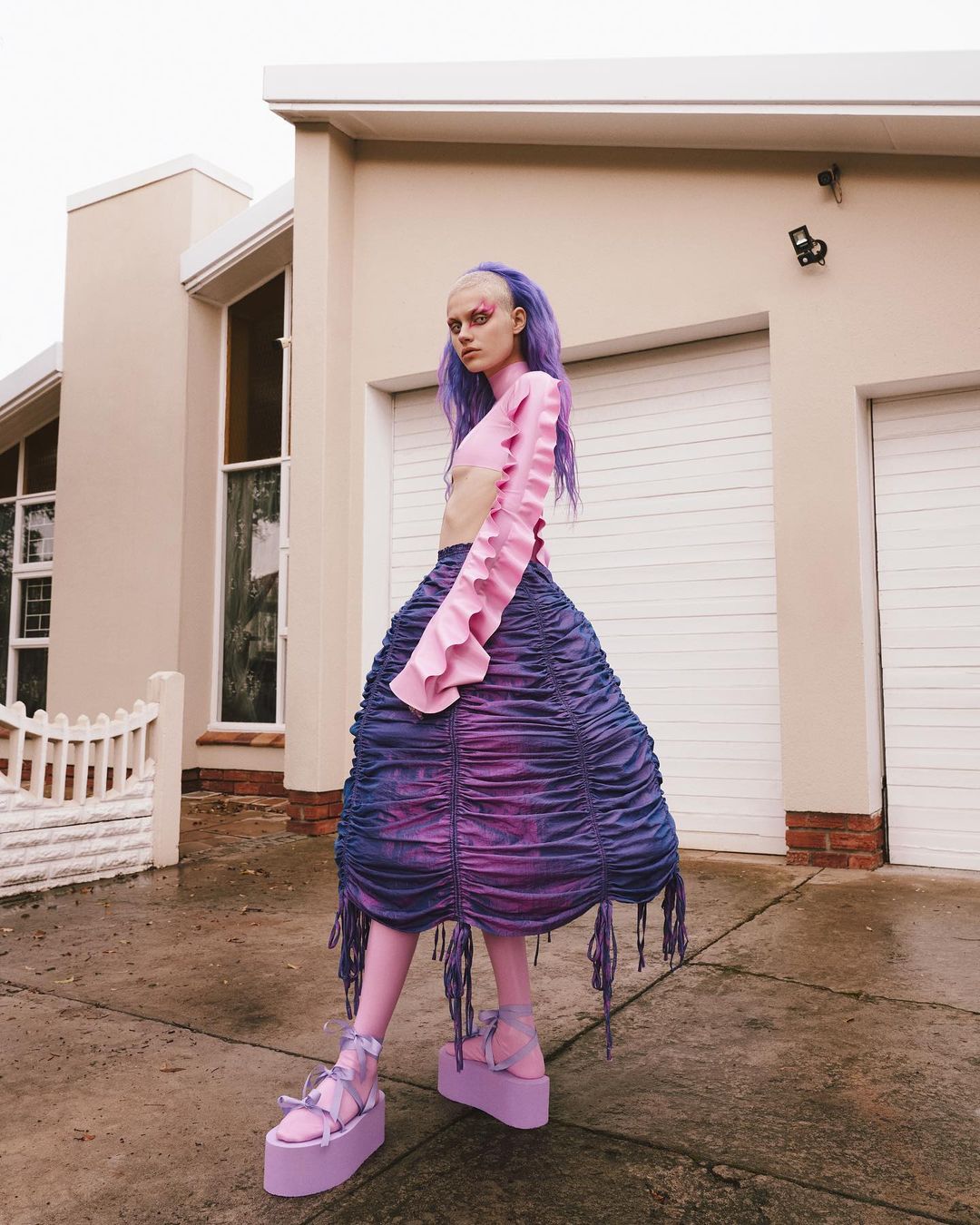 A woman with light skin and purple hair poses outside a cream-colored house wearing a purple and pink skirt and top with platform shoes.