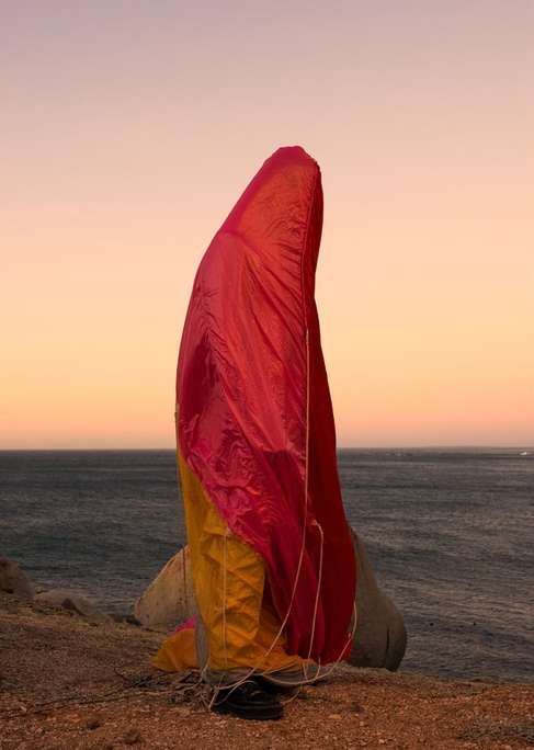 Paragliding canopy resting on a rocky outcrop near the ocean under a gradient sky at dusk, featuring pink, yellow, and blue hues.