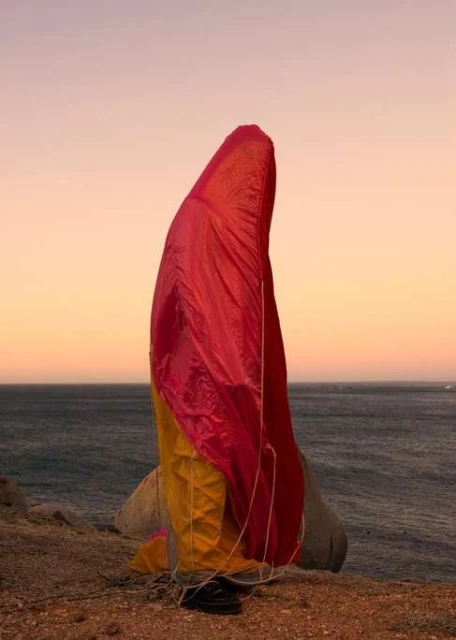 Paragliding canopy resting on a rocky outcrop near the ocean under a gradient sky at dusk, featuring pink, yellow, and blue hues.