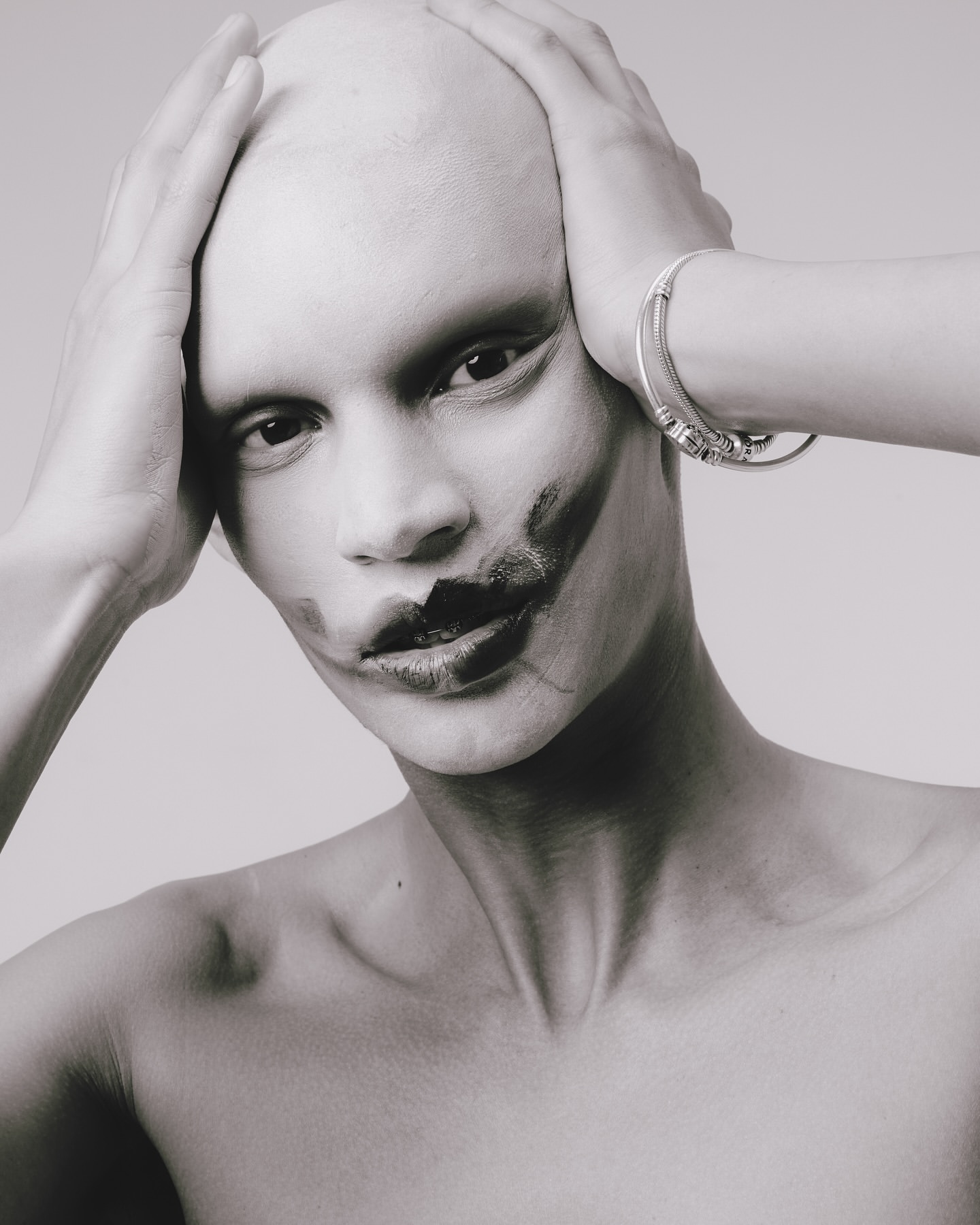 Close-up black and white portrait of a bald adult with dark makeup on their face and wearing a silver bracelet against light gray backdrop.