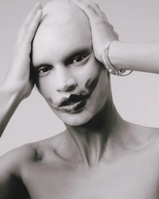 Close-up black and white portrait of a bald adult with dark makeup on their face and wearing a silver bracelet against light gray backdrop.