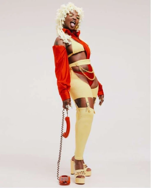 Stylish woman in red and yellow outfit, platform heels, and platinum hair strikes a pose while holding a red rotary phone.