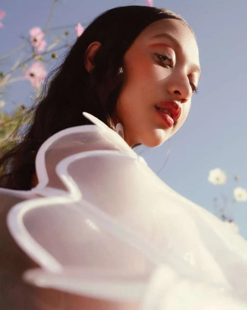 A woman with dark hair poses in a low-angle shot with a sheer white garment and pink cosmos flowers in the background against a bright blue sky.