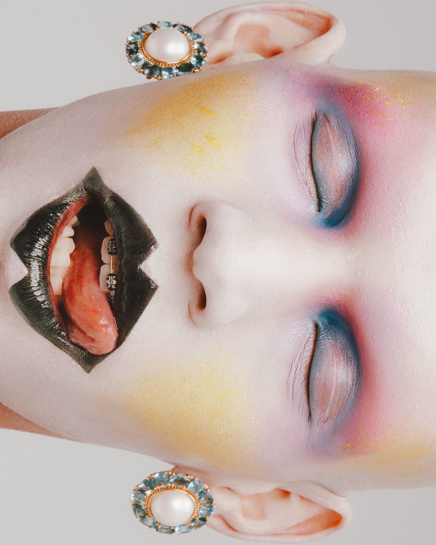 Artistic beauty portrait of a woman with colorful makeup, braces, tongue, and pearl earrings against a neutral gradient backdrop.