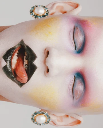 Artistic beauty portrait of a woman with colorful makeup, braces, tongue, and pearl earrings against a neutral gradient backdrop.