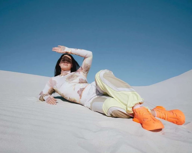 Woman in patterned top and colorful pants poses in a desert landscape with bright orange sneakers under a clear blue sky.