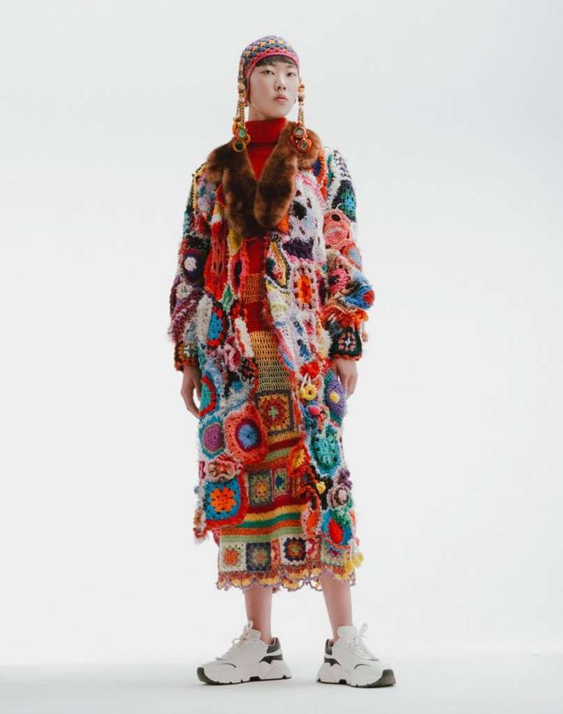 Asian model in vibrant, colorful, patchwork coat with fur collar and headdress, posing against a plain white background in a studio.