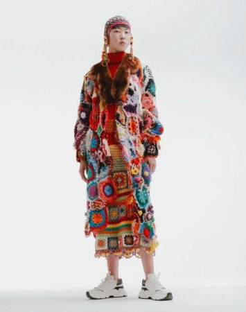 Asian model in vibrant, colorful, patchwork coat with fur collar and headdress, posing against a plain white background in a studio.