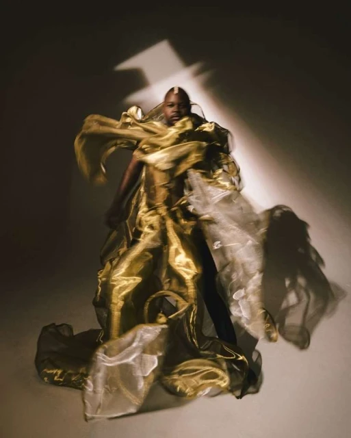 Woman poses in a shimmering gold gown in a studio with dramatic lighting, exuding avant-garde fashion in a full-length shot.