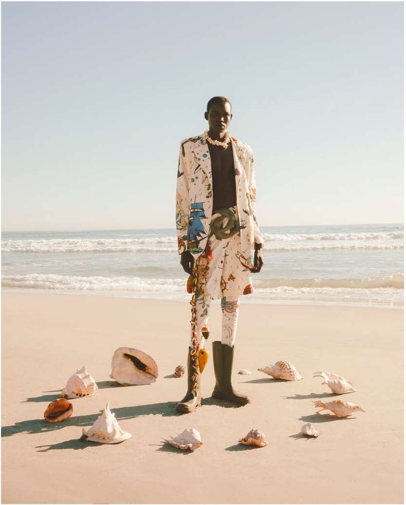 An adult African-American male poses on a beach wearing a patterned suit and boots, surrounded by seashells arranged in a circle.
