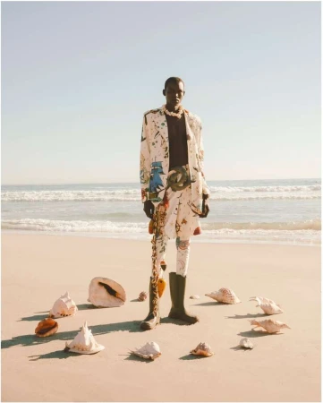 An adult African-American male poses on a beach wearing a patterned suit and boots, surrounded by seashells arranged in a circle.