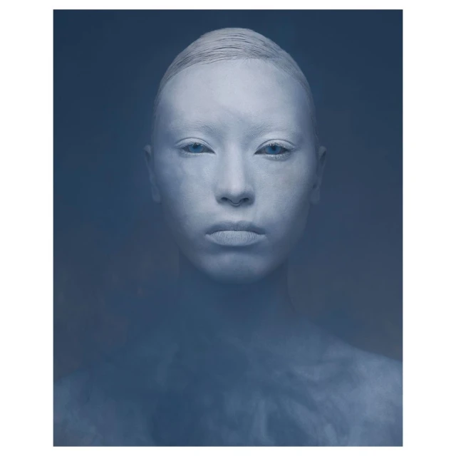 Dramatic close-up of a woman with blue eyes and white paint covering her face and hair against a dark blue background in a studio.