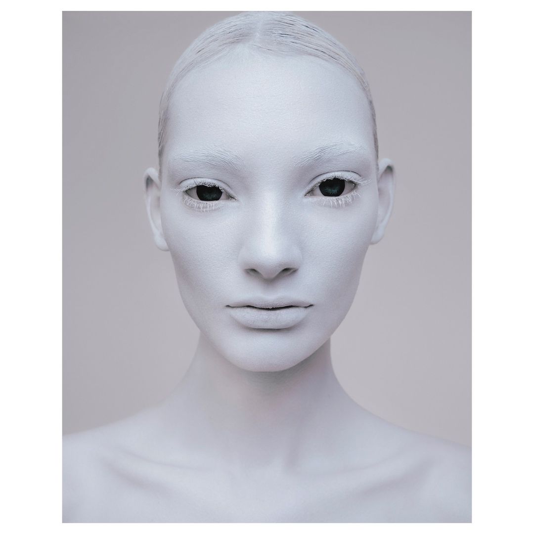 Close-up studio portrait of a woman with her face painted white and eyes black, neutral facial expression, and light gray background.