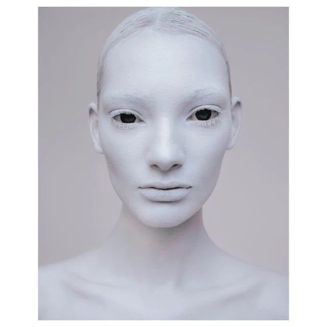 Close-up studio portrait of a woman with her face painted white and eyes black, neutral facial expression, and light gray background.