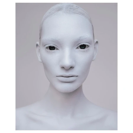 Close-up studio portrait of a woman with her face painted white and eyes black, neutral facial expression, and light gray background.