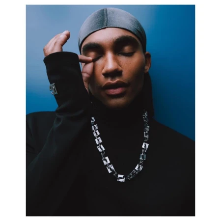 Close-up portrait of a Black man wearing a black turtleneck, silver chain necklace, and grey du-rag against a blue background.